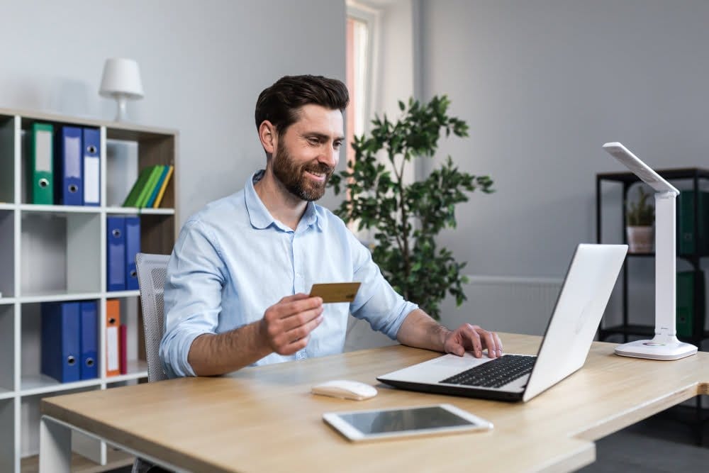 A man sitting at a desk, using a laptop and holding a credit card for online transactions.