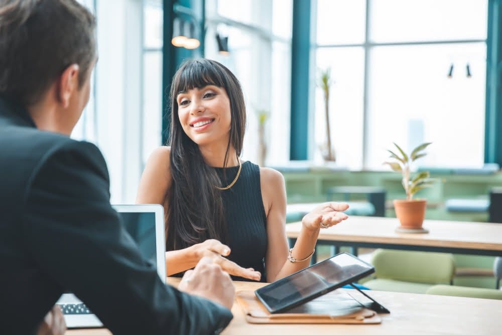 A man and woman discussing in an office, engaged in a conversation while sitting at a desk.