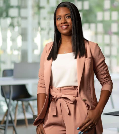 A professional woman in a pink suit confidently stands in a well-lit office space.