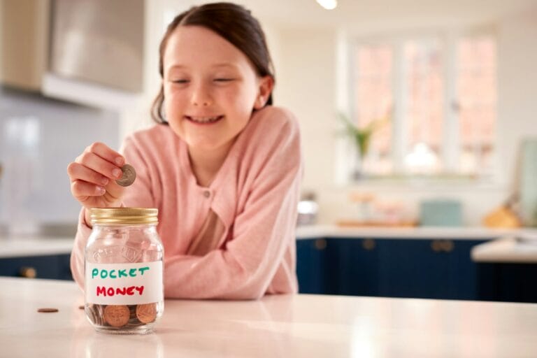 Girl Saving Money Into Jar Labelled Pocket Money On Kitchen Counter At Home