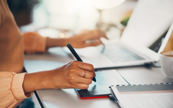 Graphic designer writing notes on tablet online, typing on laptop and planning a task working remot