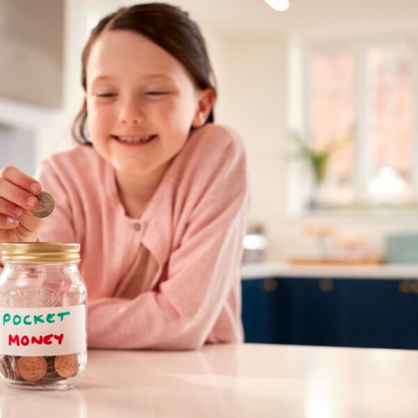 Girl Saving Money Into Jar Labelled Pocket Money On Kitchen Counter At Home